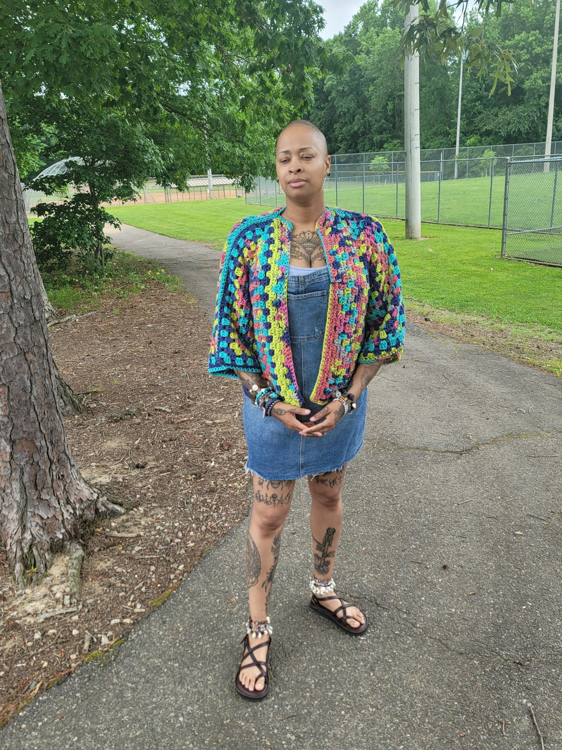 A woman is standing on a sidewalk wearing overalls and a colorful jacket.