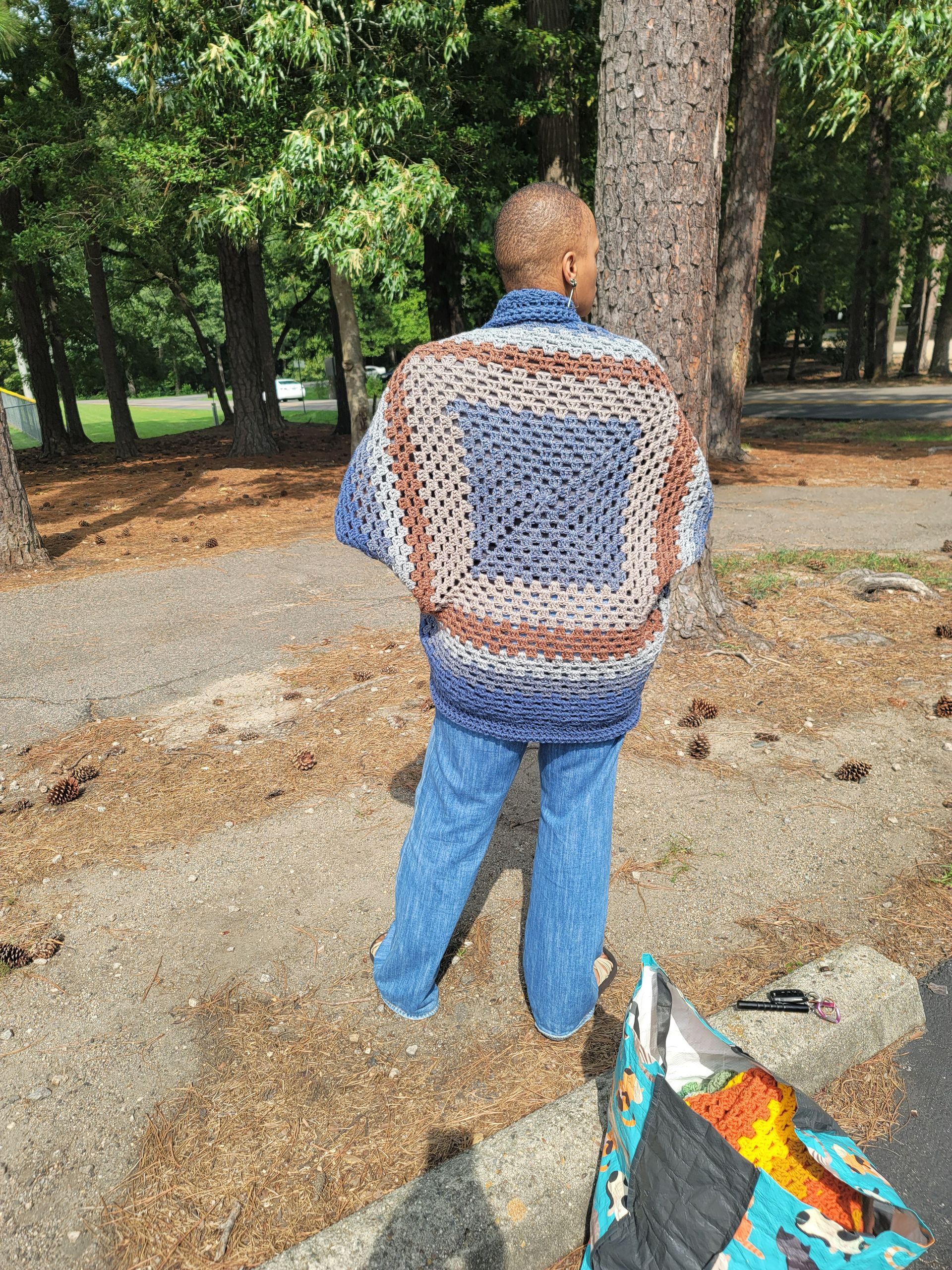 A man wearing a crocheted jacket is standing in a park.