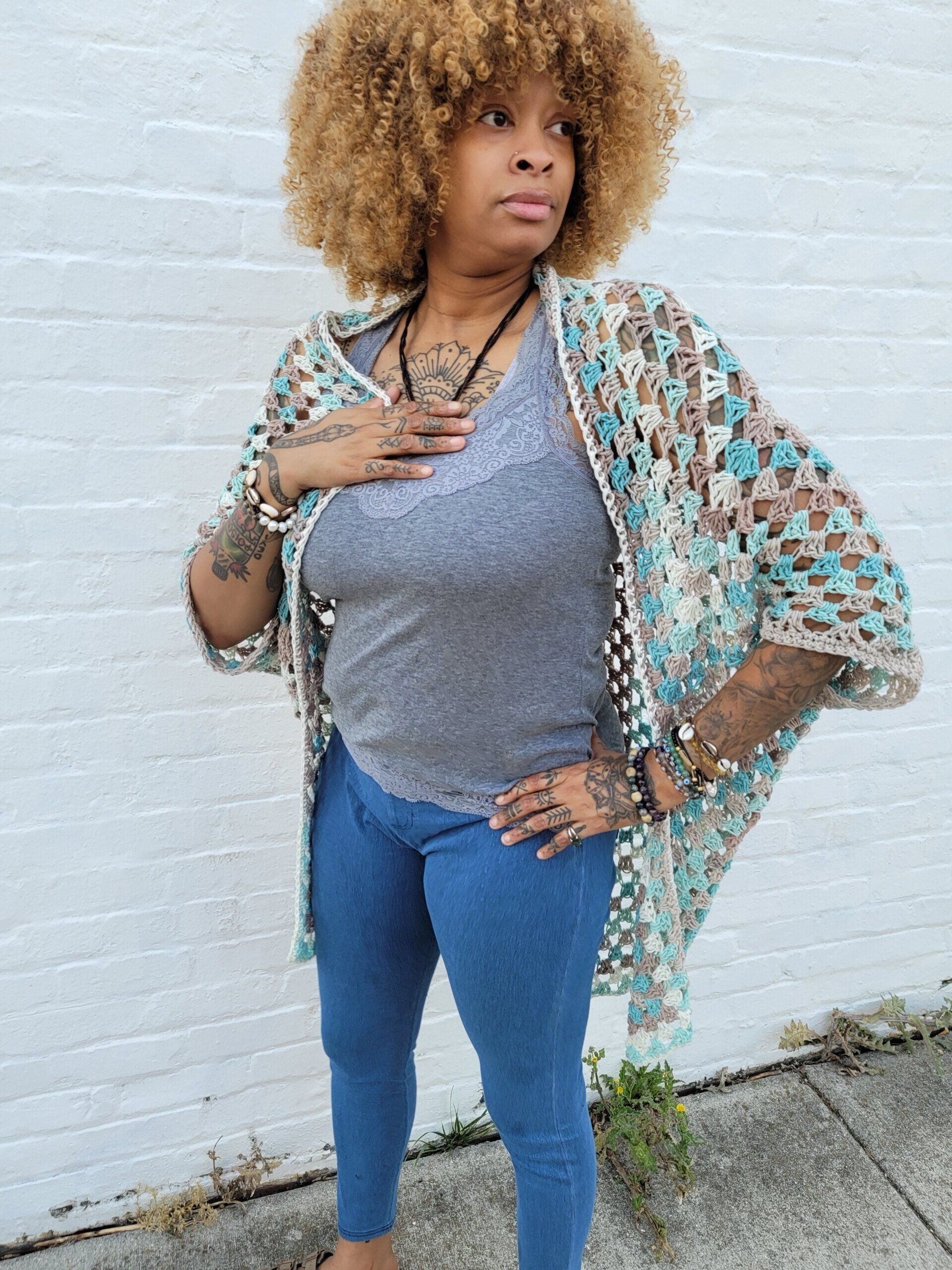 A woman wearing a crocheted cardigan and jeans is standing in front of a white brick wall.