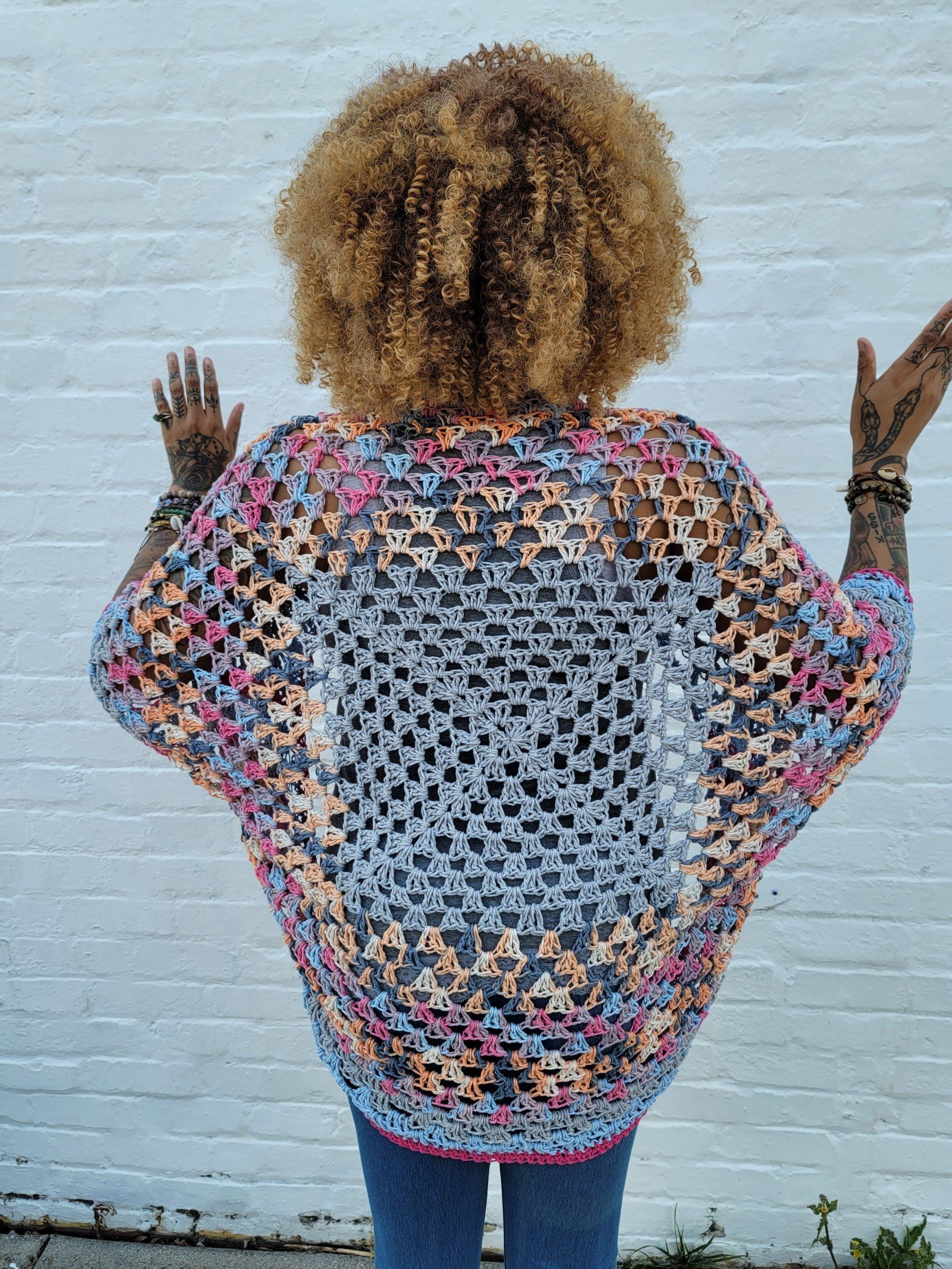 A woman is standing in front of a white brick wall wearing a crocheted sweater.