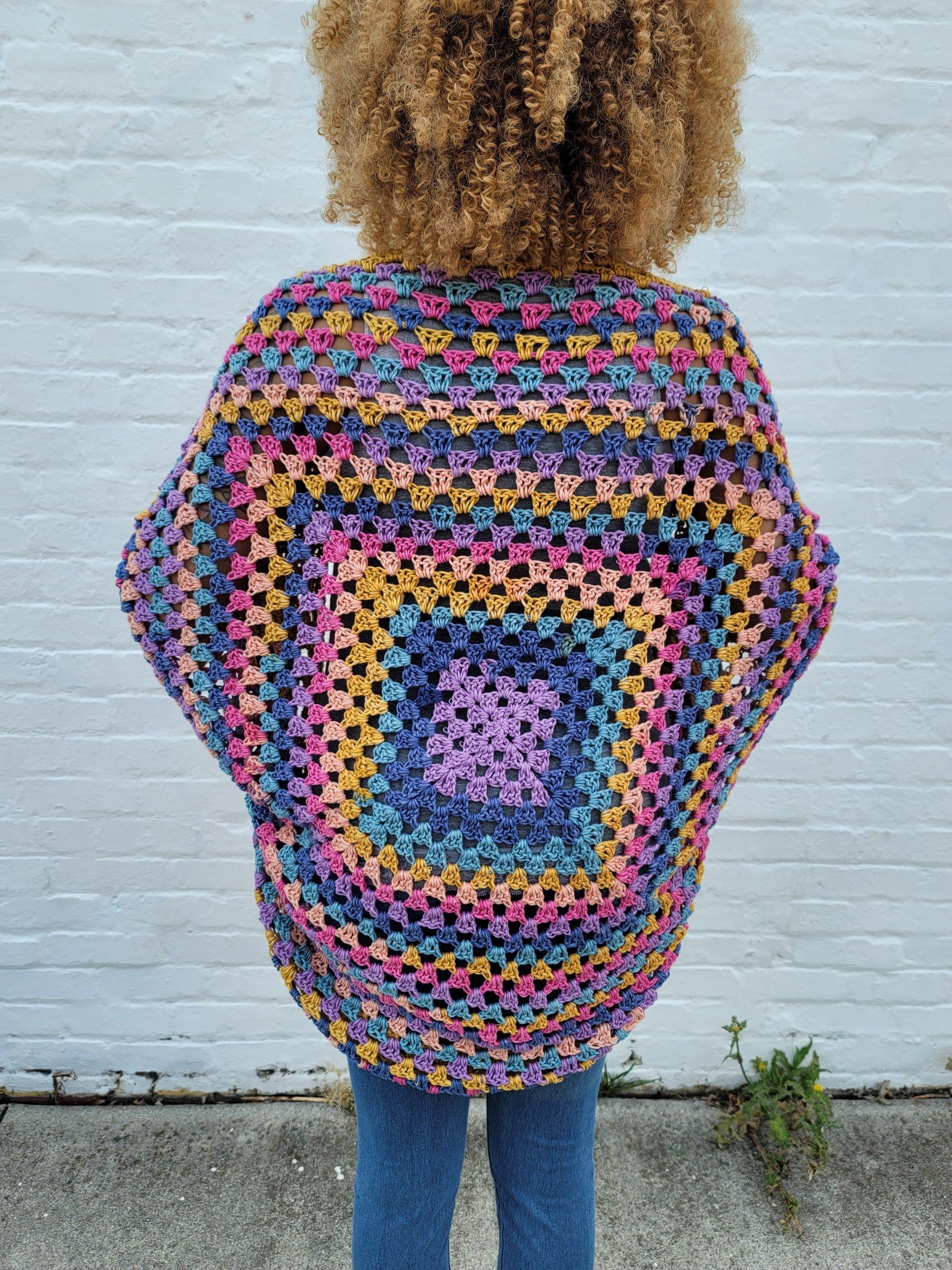 A woman wearing a colorful crocheted sweater is standing in front of a white brick wall.