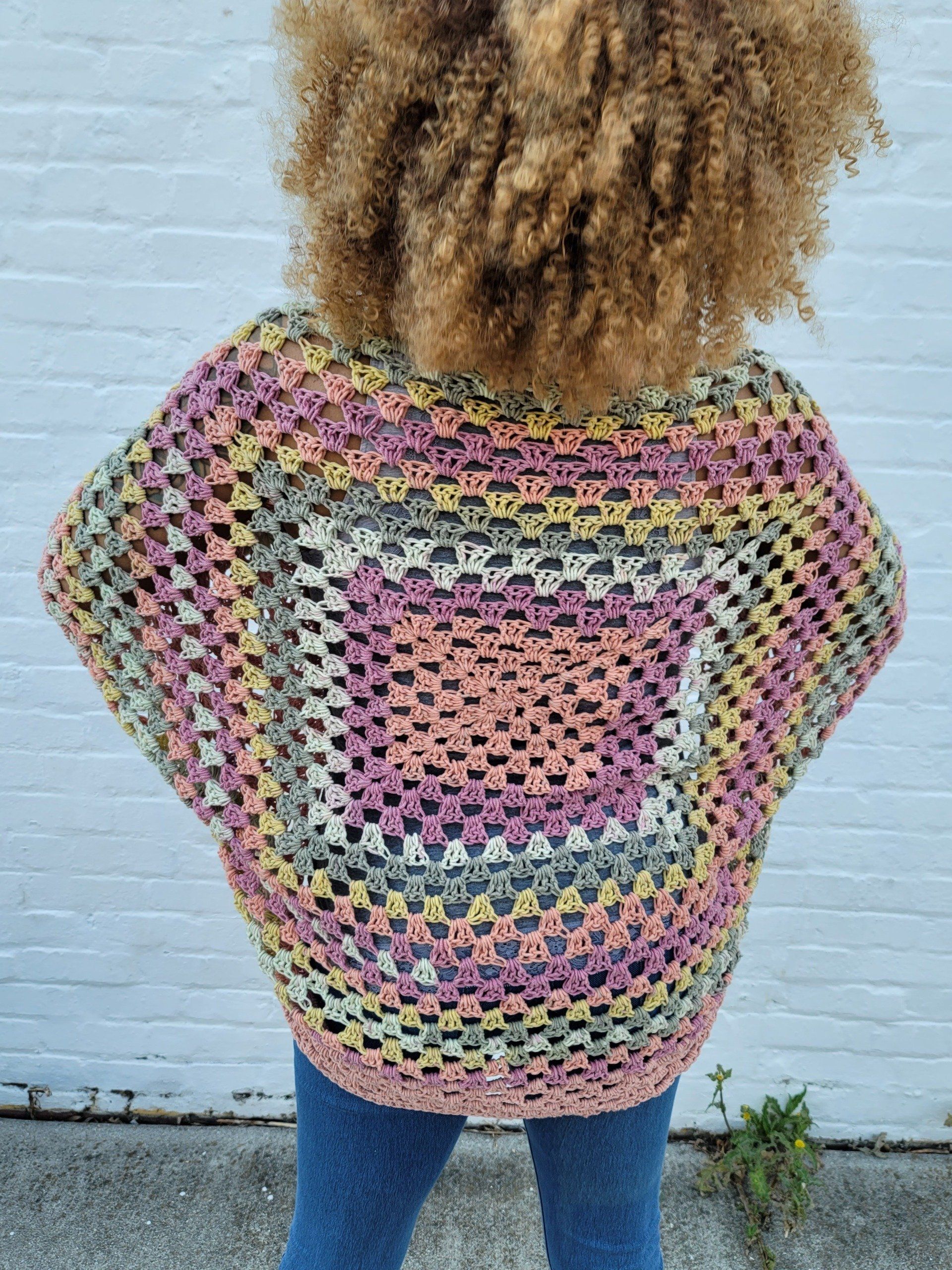 A woman is standing in front of a white brick wall wearing a colorful crocheted sweater.