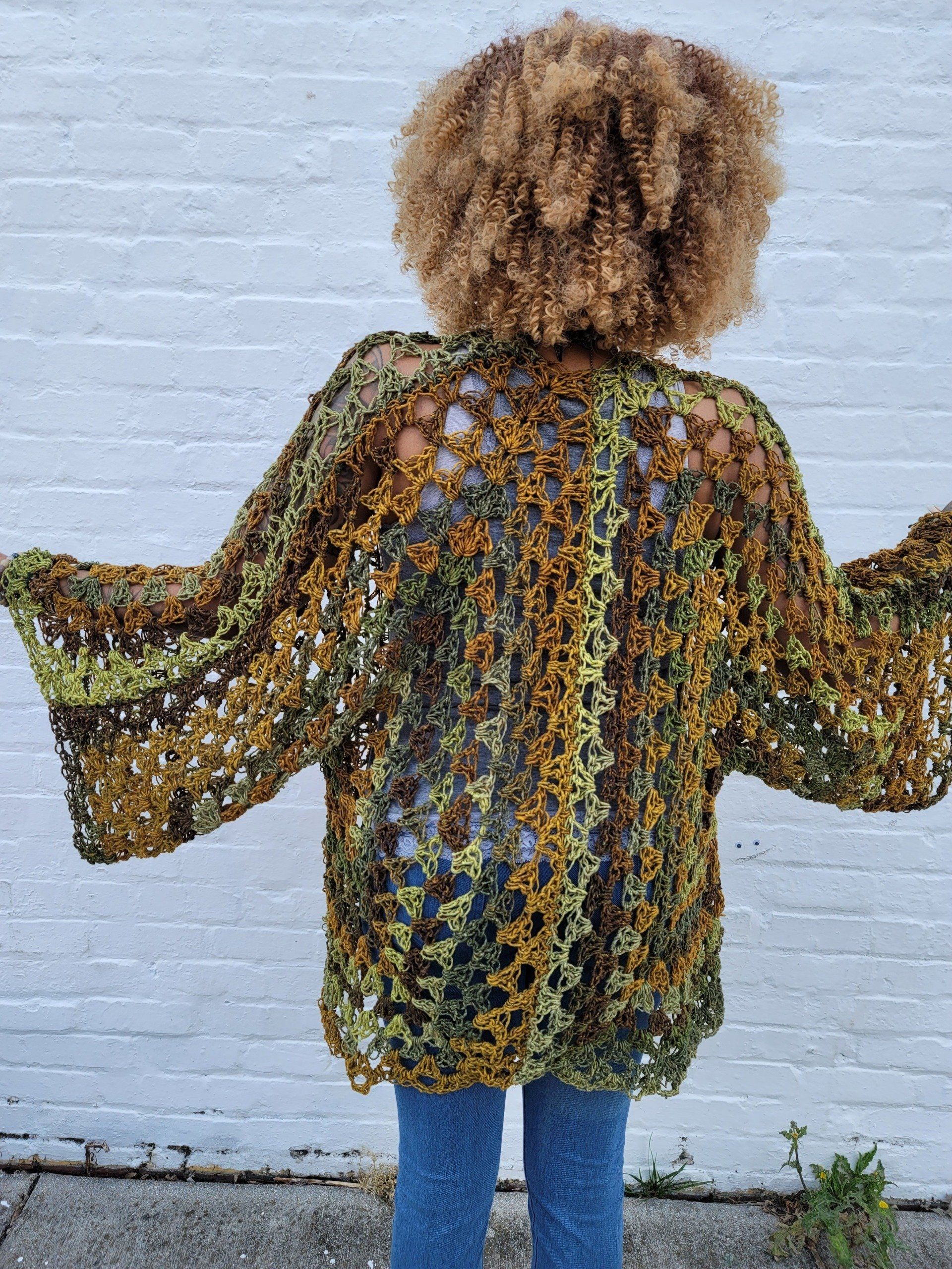 A woman wearing a crocheted jacket is standing in front of a white brick wall.
