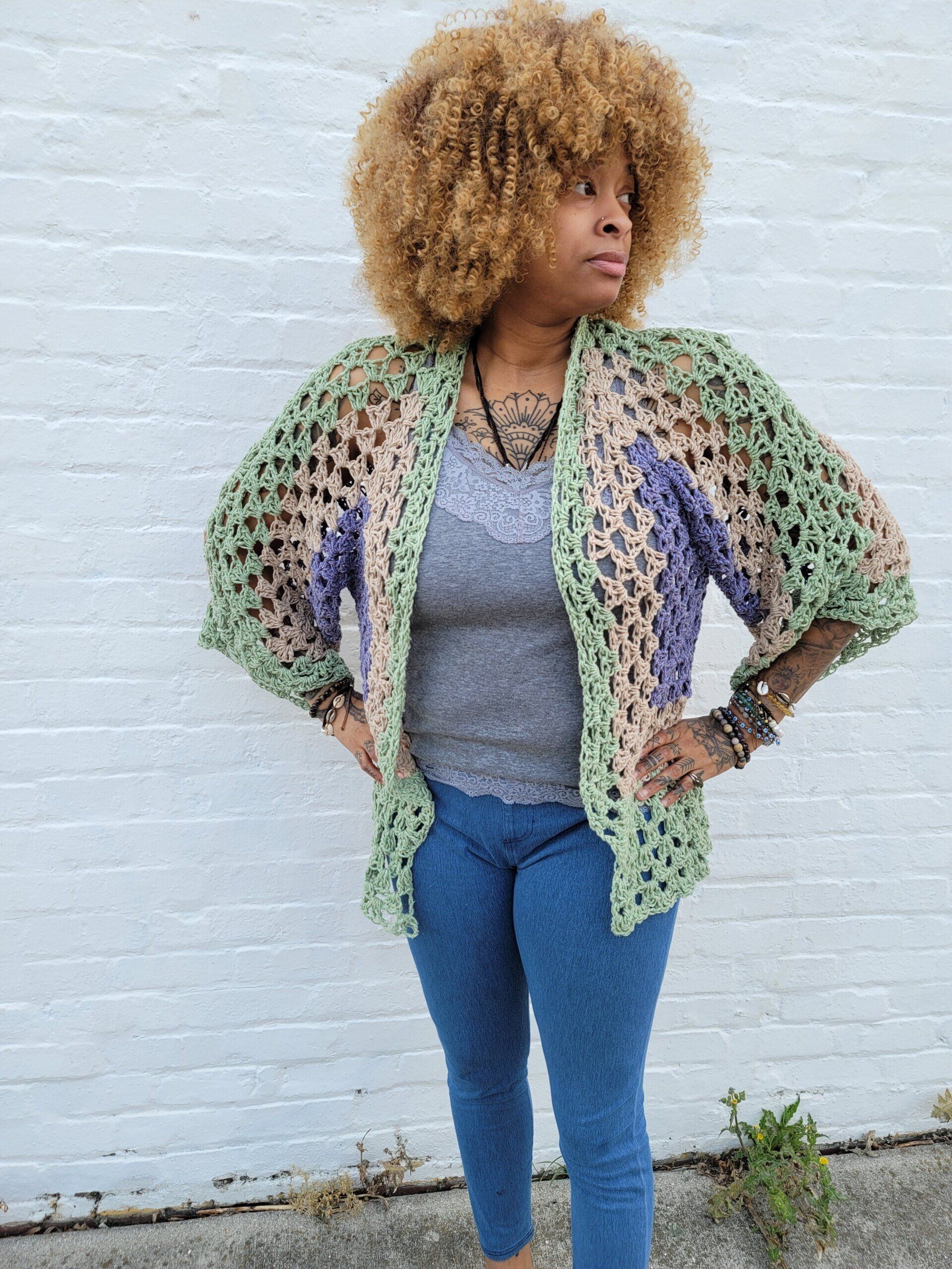 A woman wearing a crocheted cardigan is standing in front of a white brick wall.