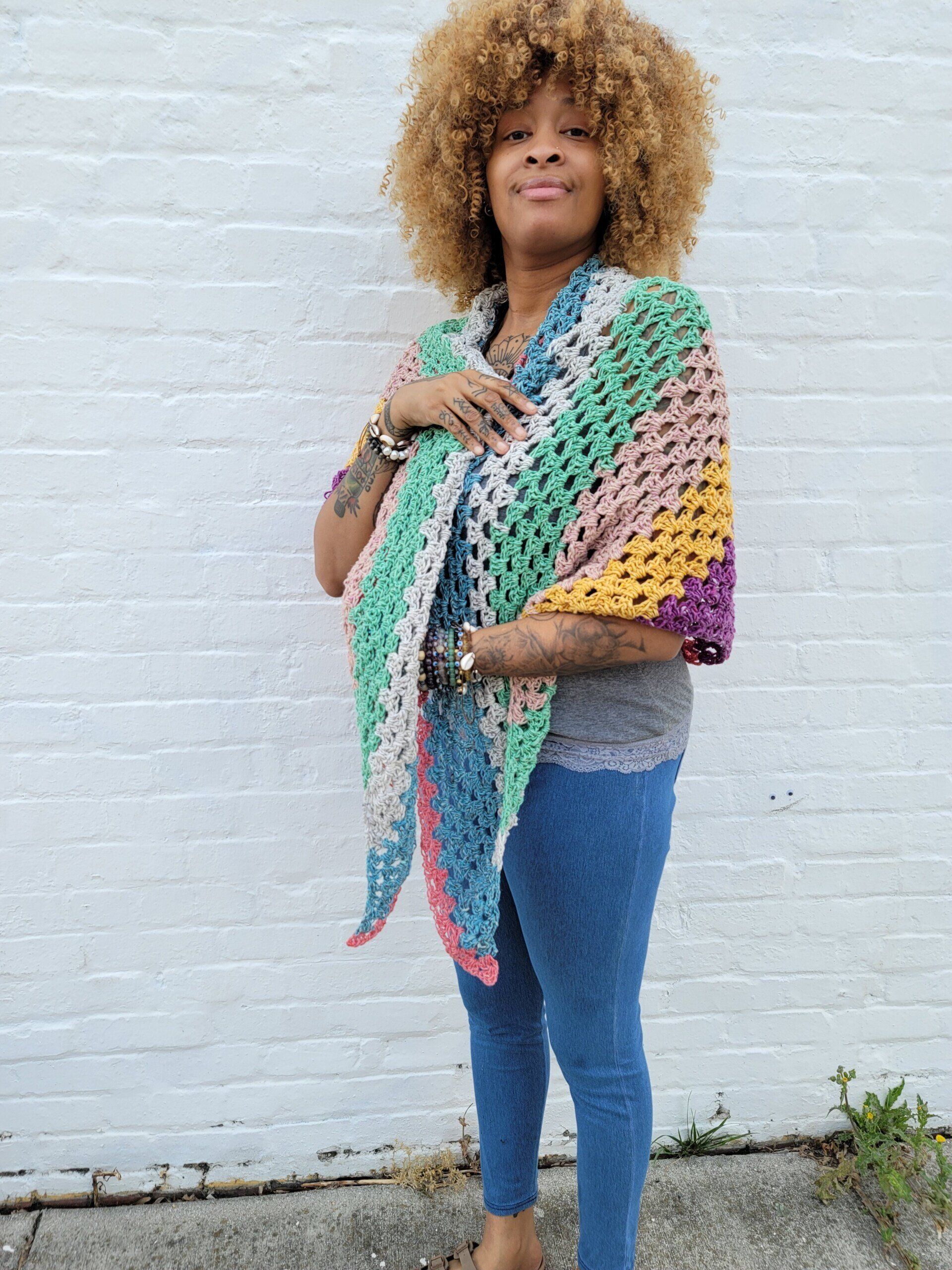 A woman wearing a colorful crocheted shawl is standing in front of a white brick wall.