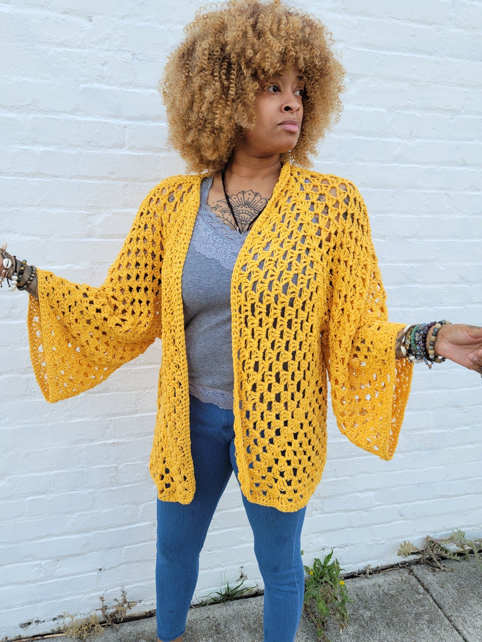 A woman wearing a yellow crocheted cardigan is standing in front of a white wall.