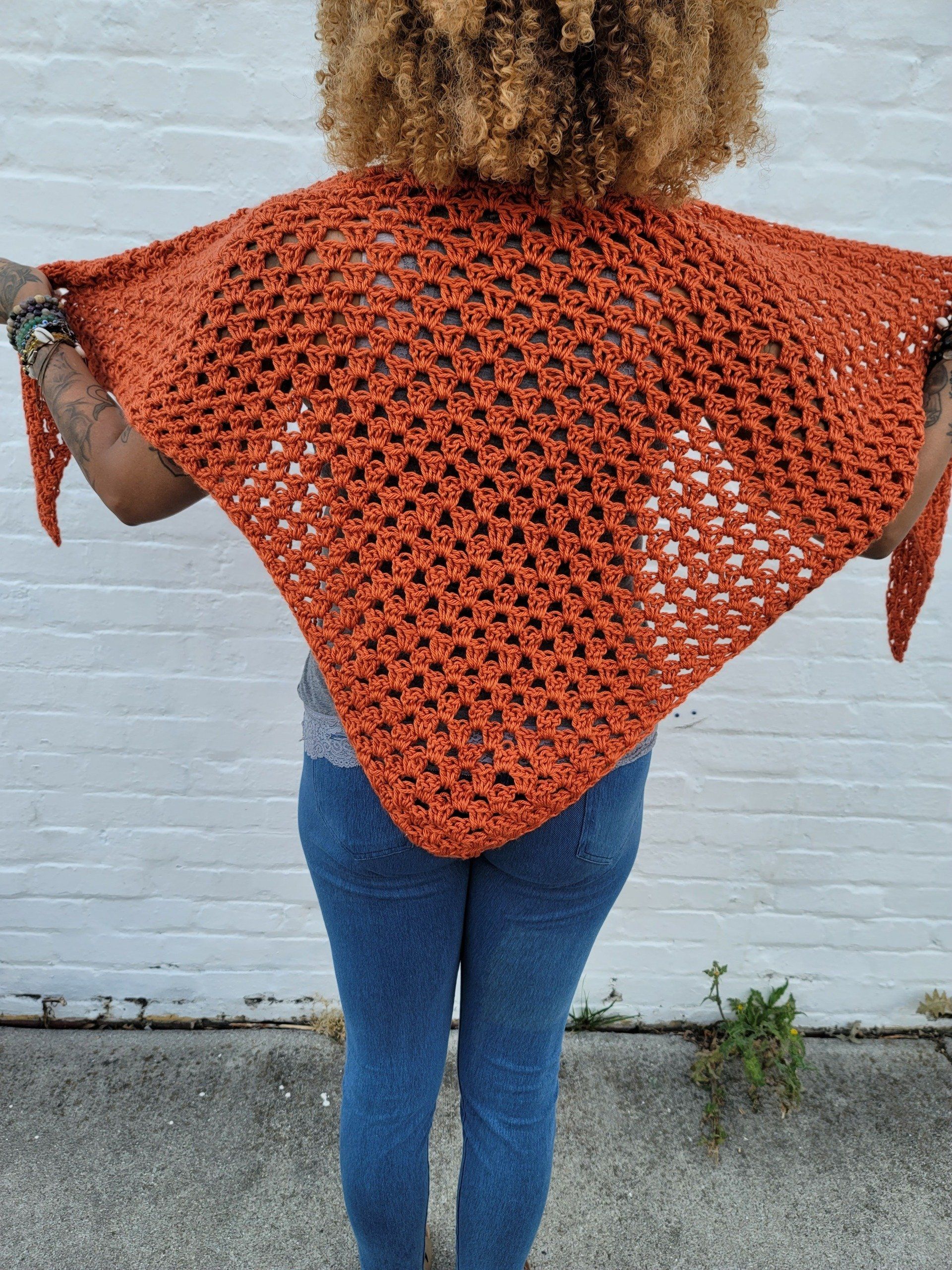 A woman wearing a crocheted shawl is standing in front of a white brick wall.