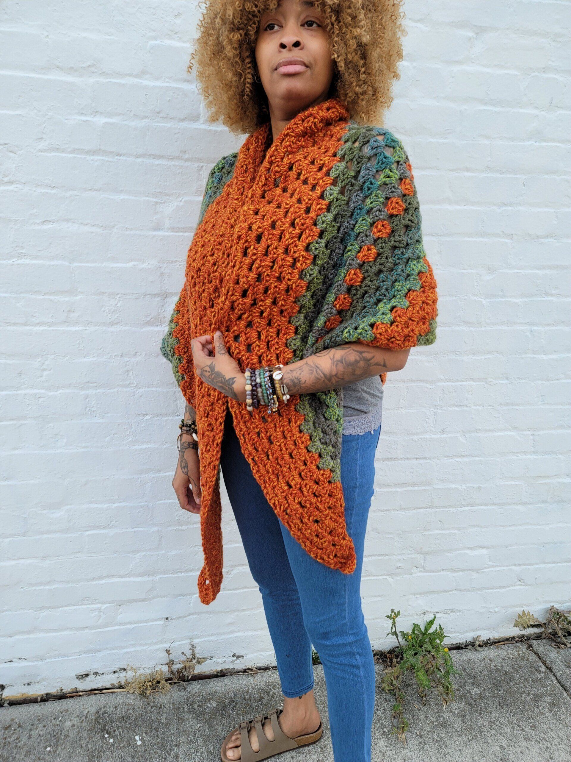 A woman wearing a crocheted shawl is standing in front of a white brick wall.