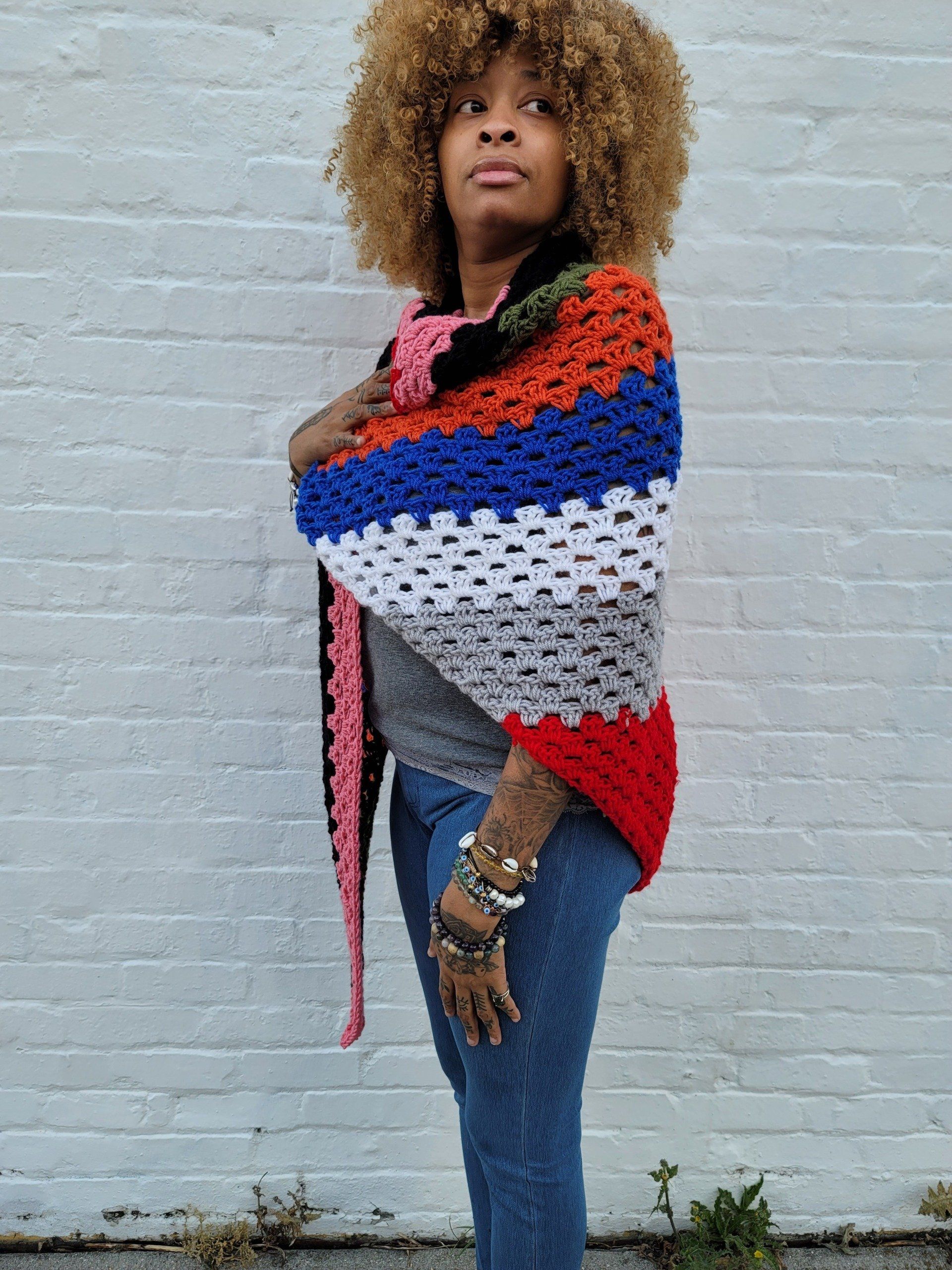 A woman wearing a colorful crocheted shawl is standing in front of a white brick wall.