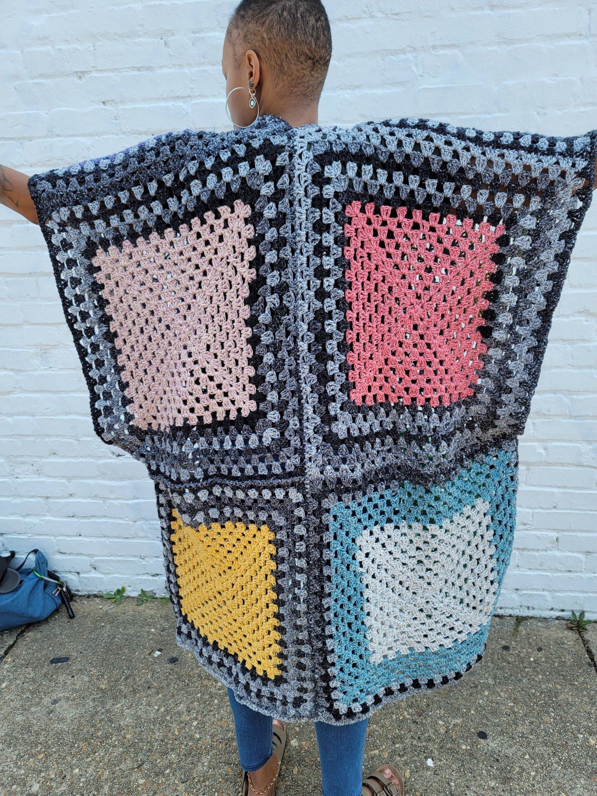 A woman is holding a crocheted blanket in front of a white brick wall.