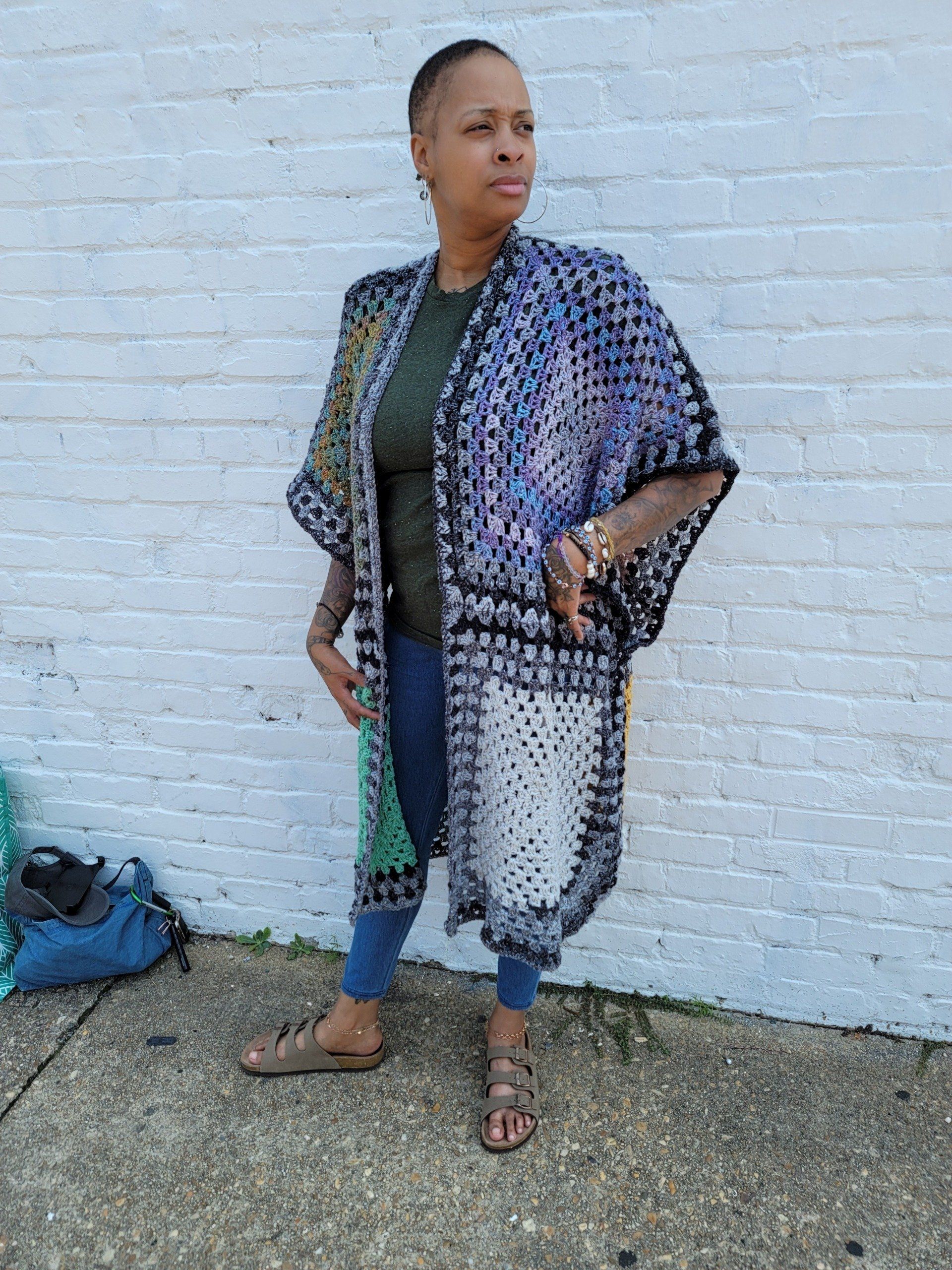 A woman wearing a crocheted kimono is standing in front of a white brick wall.