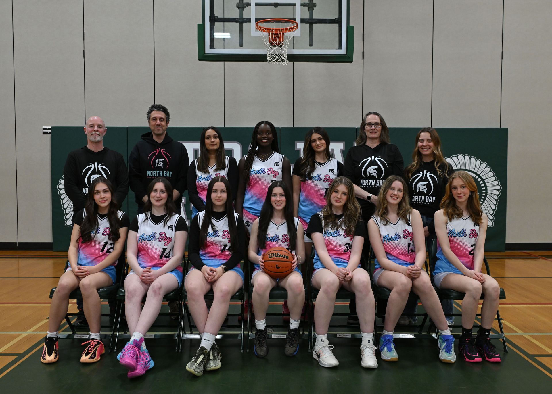 Women's basketball team in black uniforms poses, North Bay logo, some kneeling, indoors.