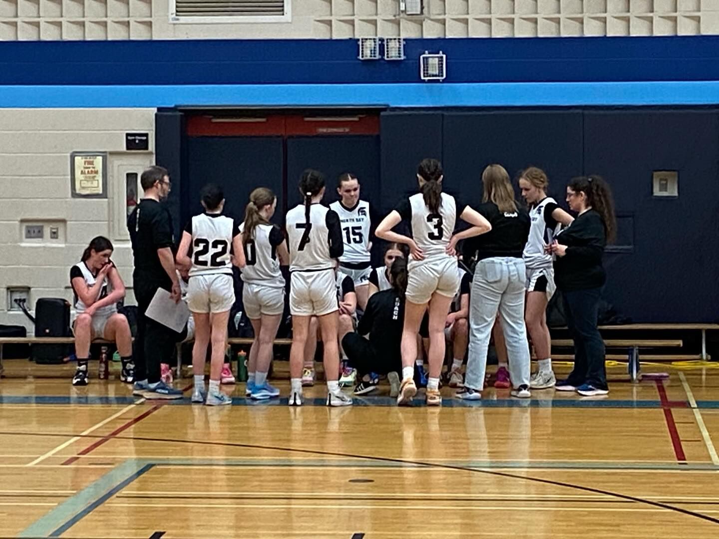 Basketball team huddle in a gymnasium. Players in black/white uniforms listen to a coach.