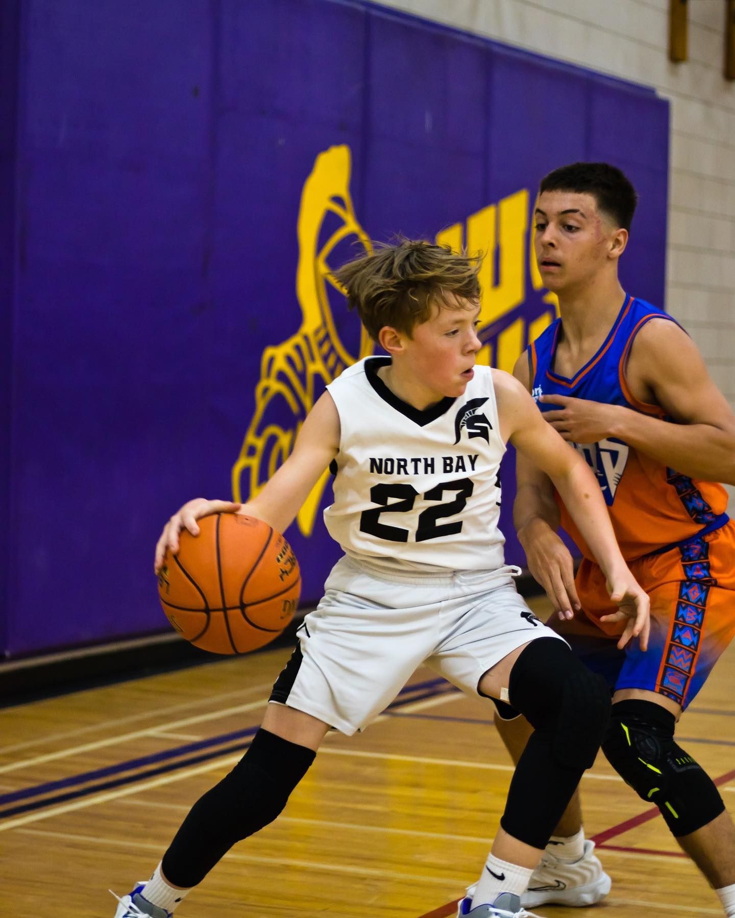 Basketball player in white uniform dribbling, defended by another player in an orange jersey. Court in background.