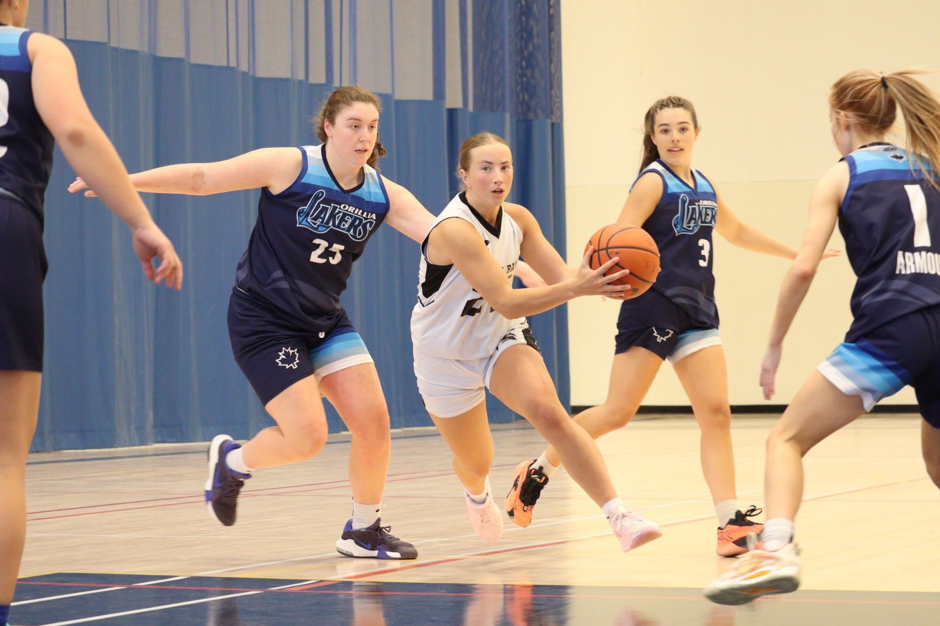 Basketball player dribbling the ball, pursued by two opponents in blue. Indoor court setting.