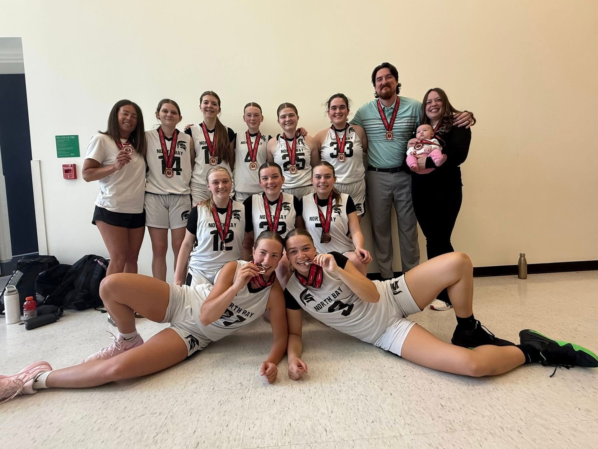 Basketball team posing with medals. Players in black and white uniforms. Smiling faces, indoors.