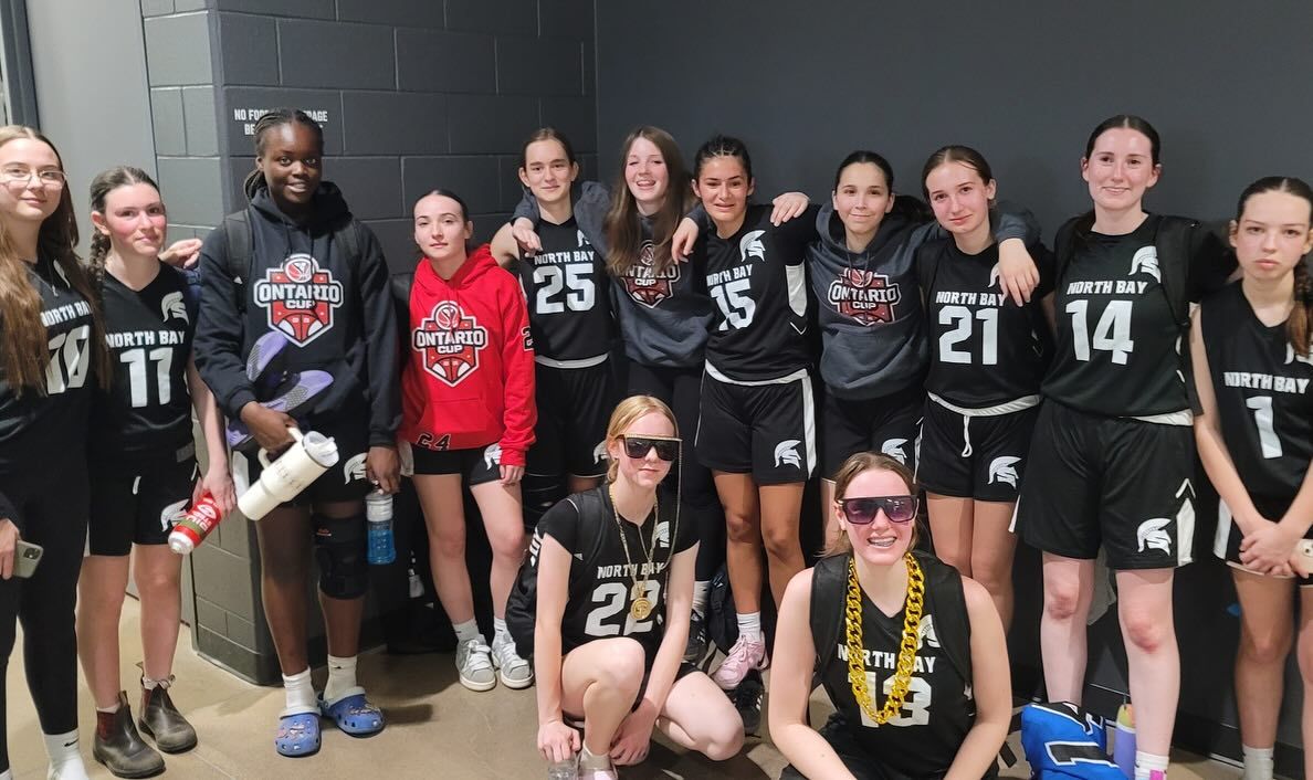 A basketball team poses in a locker room. Players wear black jerseys with white numbers, some with sunglasses.