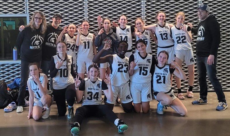 Women's basketball team poses for photo indoors, smiling and gesturing; team is wearing black and white jerseys.