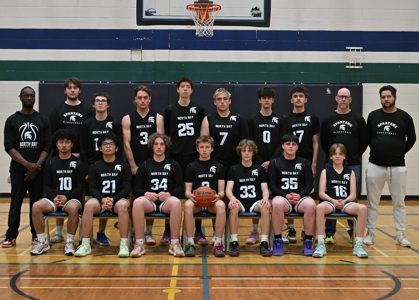 Basketball team portrait, black jerseys, gymnasium setting. Players seated, some holding a basketball, with coaches.