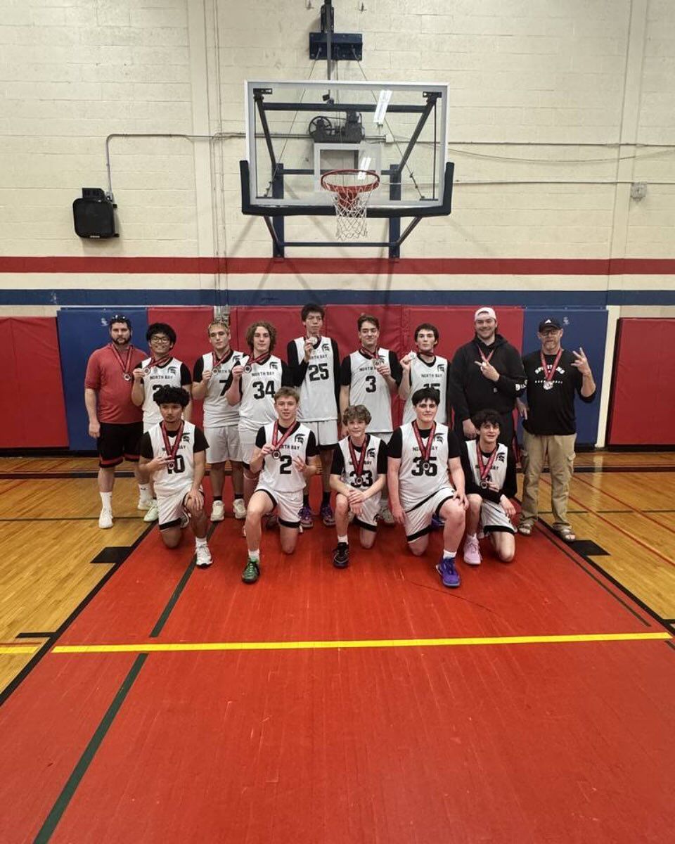 Basketball team, kneeling and standing on court, posing after winning; wearing medals, black and white uniforms, red and blue backdrop.
