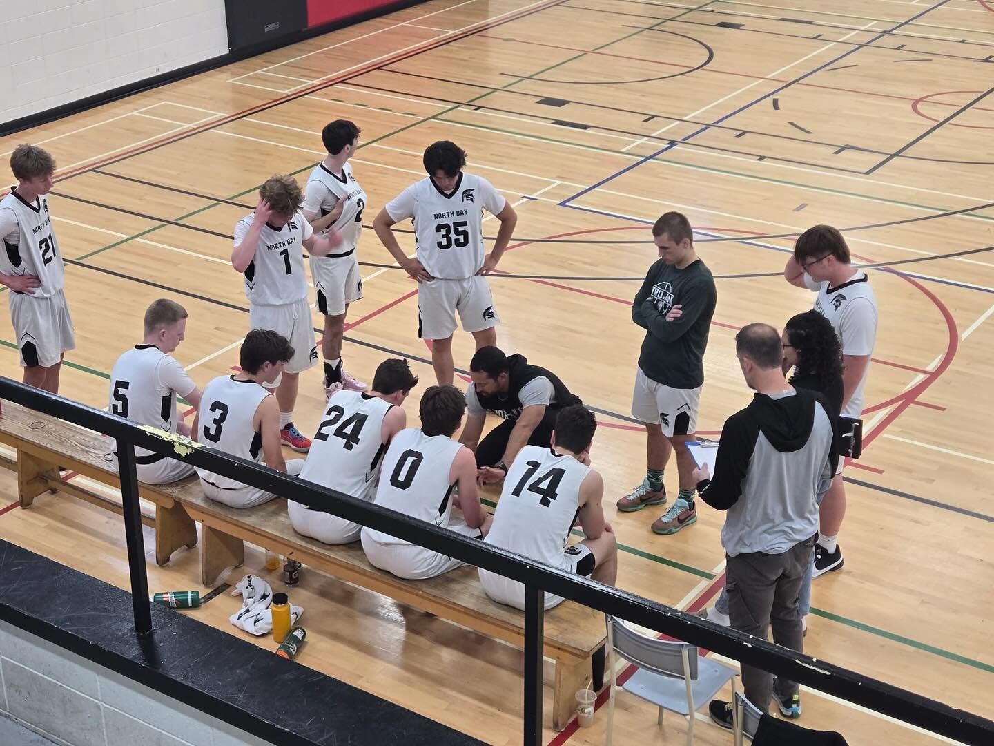 Basketball team huddle on court; coach talks strategy. Players wear white jerseys, one with 