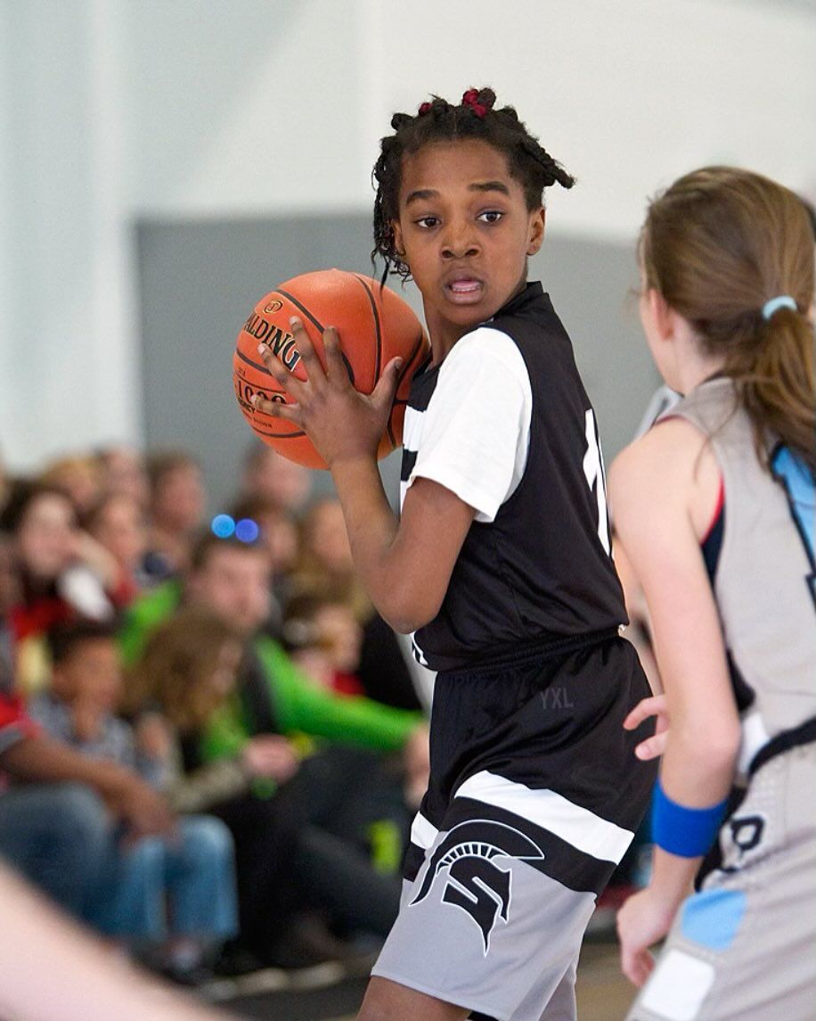 Girl in black basketball uniform dribbles the ball on court, looking intently at a competitor.