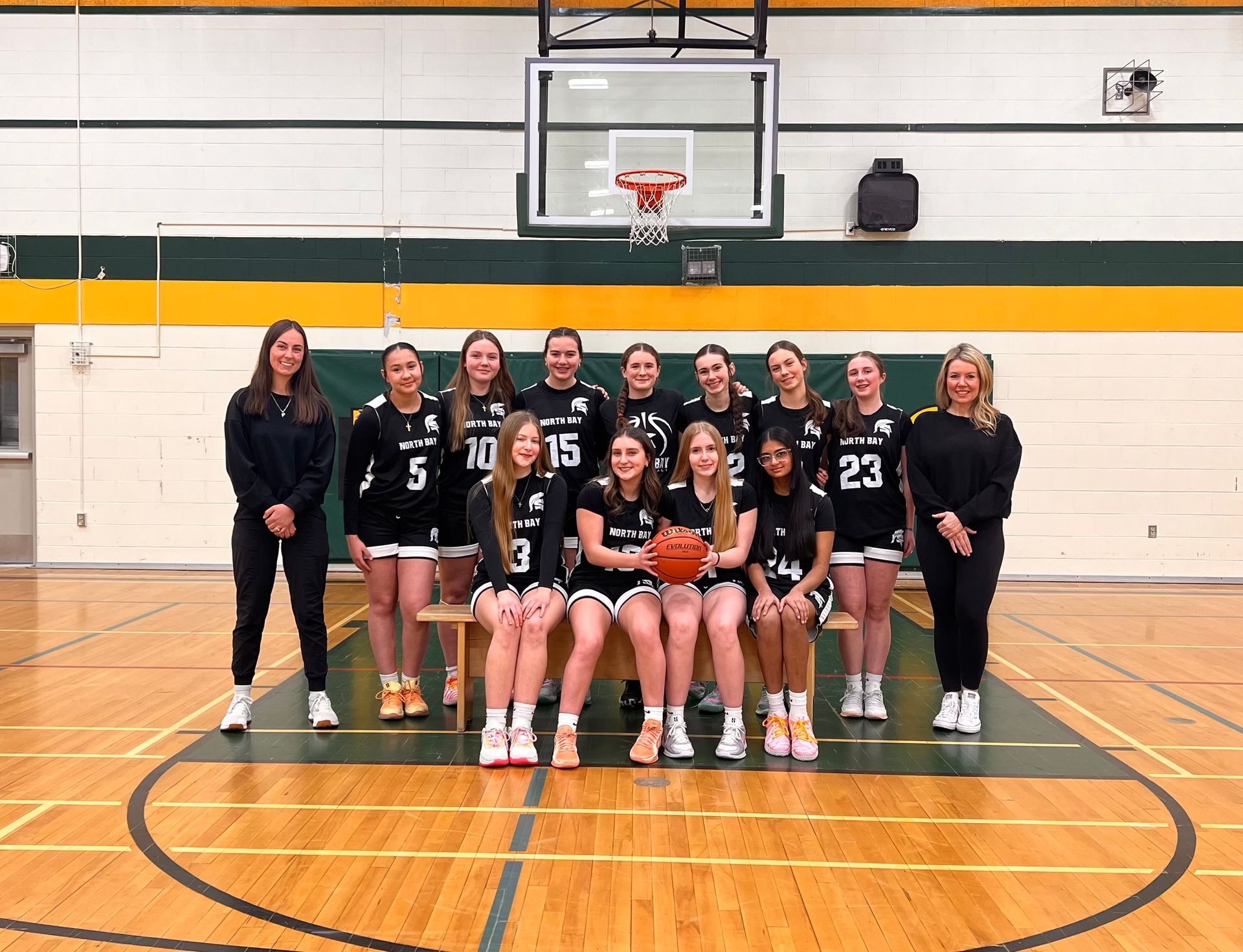Women's basketball team poses. Black and white uniforms, gesturing, indoors in front of a patterned wall.