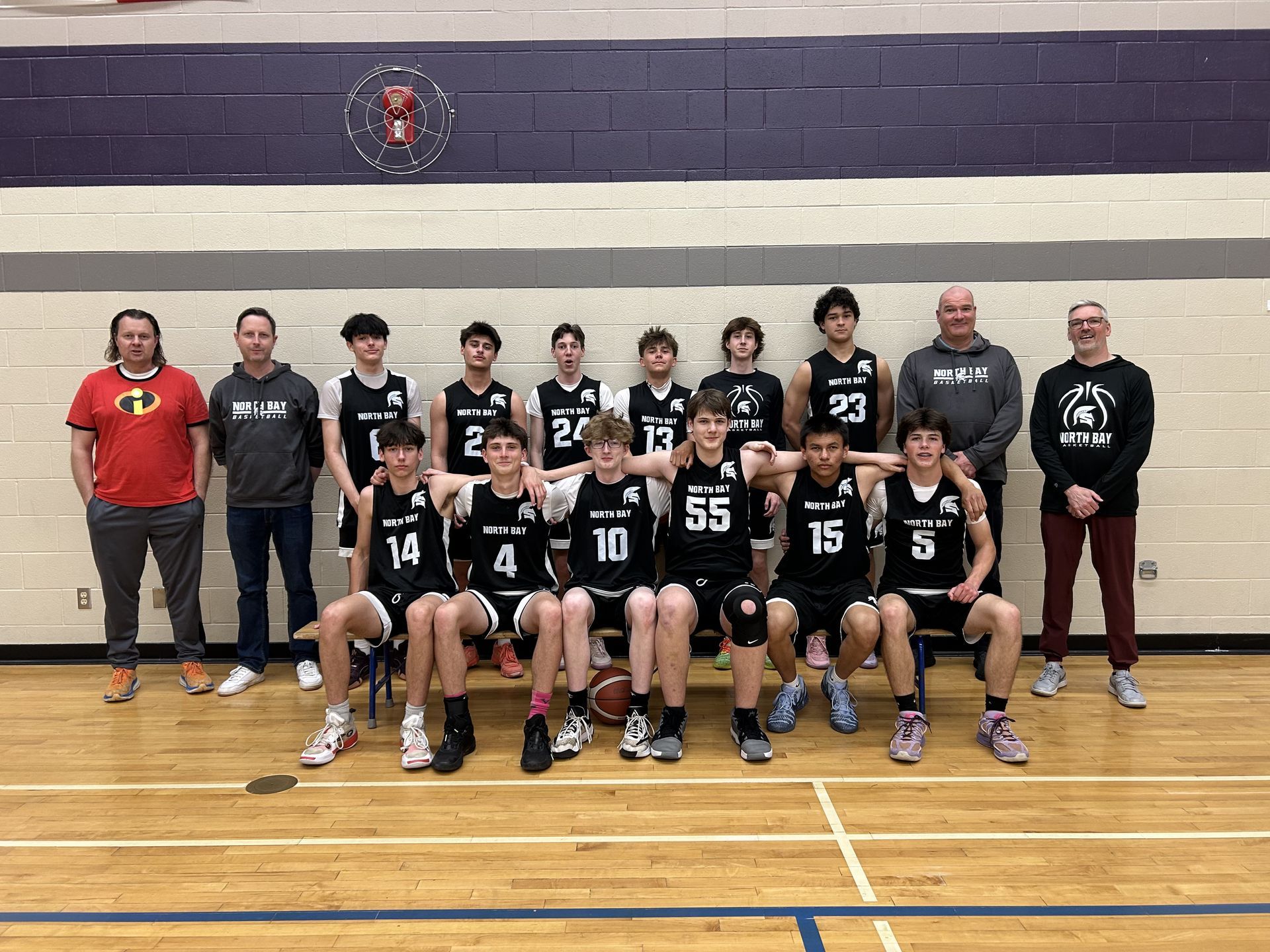Basketball team photo: group of teenage boys in black jerseys with coaches, posed indoors.