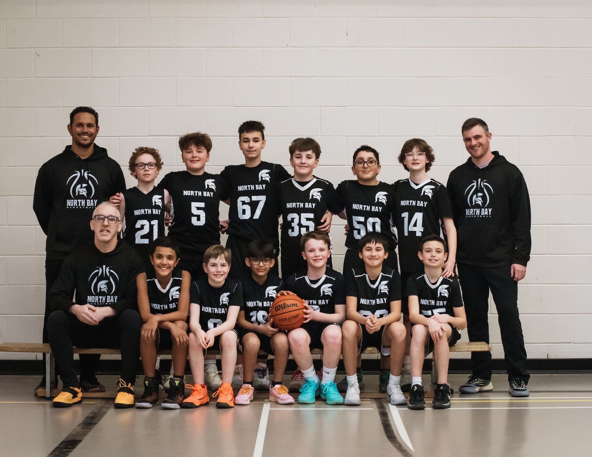 Basketball team, white uniforms, posing on a court. Players & coaches smiling, some holding basketball.