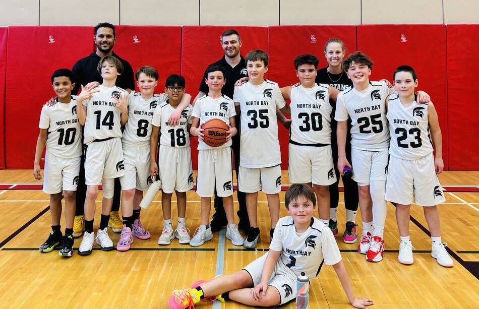 Basketball team, white uniforms, posing on a court. Players & coaches smiling, some holding basketball.