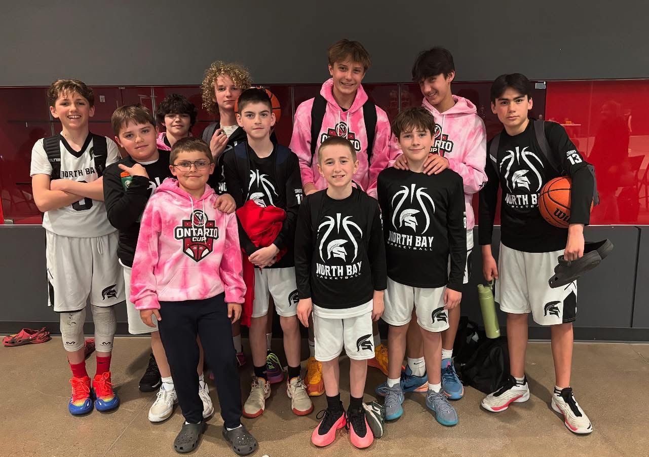 Basketball team poses together. Boys in black & pink outfits, some holding basketballs, in a red-walled area.