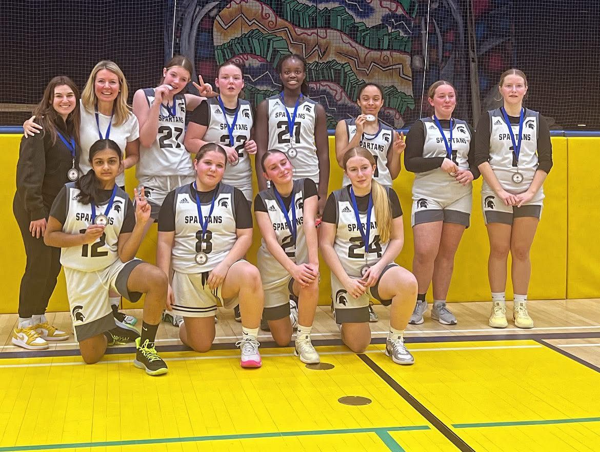 Basketball team of young women wearing silver medals, posing on a yellow court.