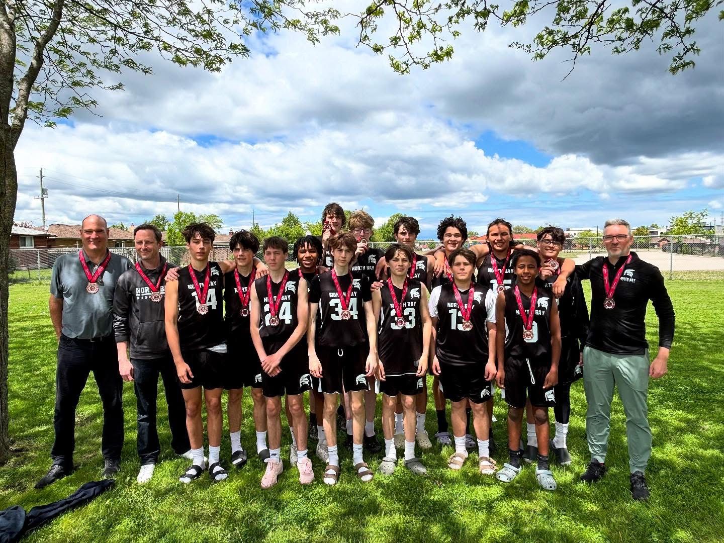 A youth basketball team in black jerseys with medals poses for a photo on a grassy field under a blue sky.