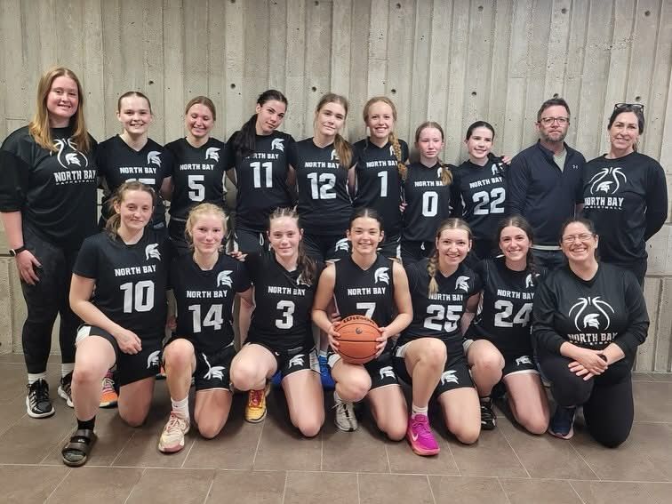Women's basketball team in black uniforms poses, North Bay logo, some kneeling, indoors.