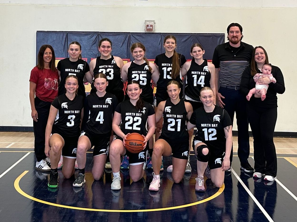 Basketball team poses for photo, black jerseys, some kneeling. Coaches and baby on the side.