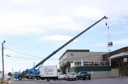 A blue crane lifting an HVAC unit onto a building. A truck and parked cars are visible. Sunny day.
