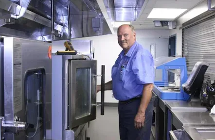 Man in blue uniform opens oven door in a stainless steel kitchen.