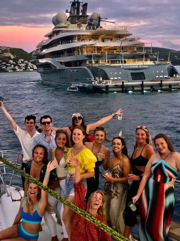 Guests leaning on the bow railing of Sailβ―Theβ―Phoenix at sunset with another yacht anchored in the distance
