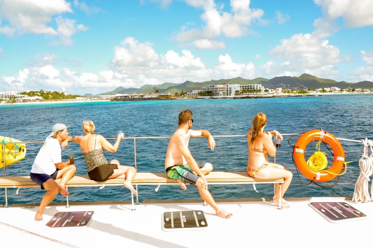 Four guests relaxing on the bow bench of Sailβ―Theβ―Phoenix as it sails along the Sintβ―Maarten coastline