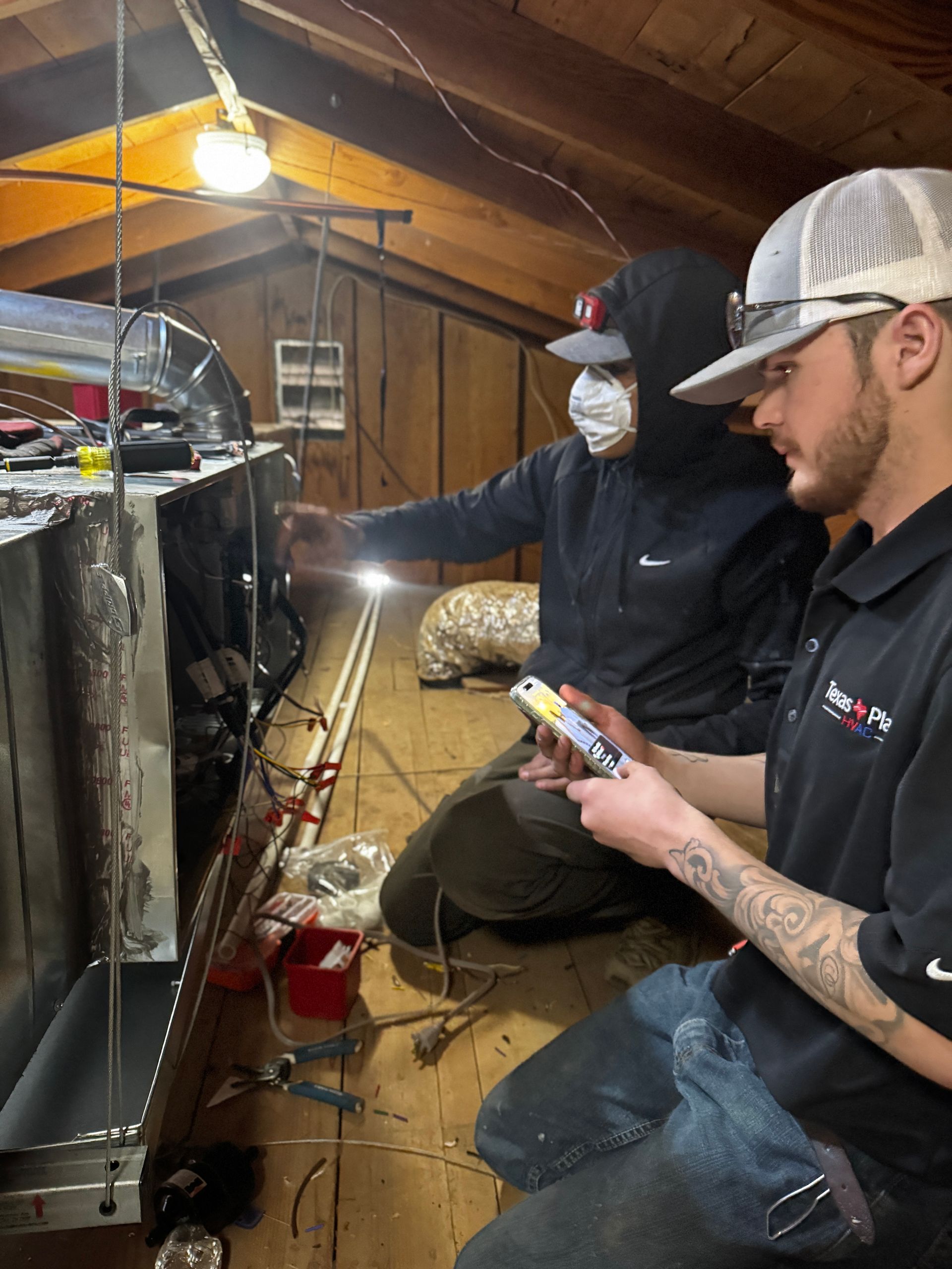 Two technicians work on an HVAC unit in an attic, one checking a handheld device and the other wearing a respirator mask.