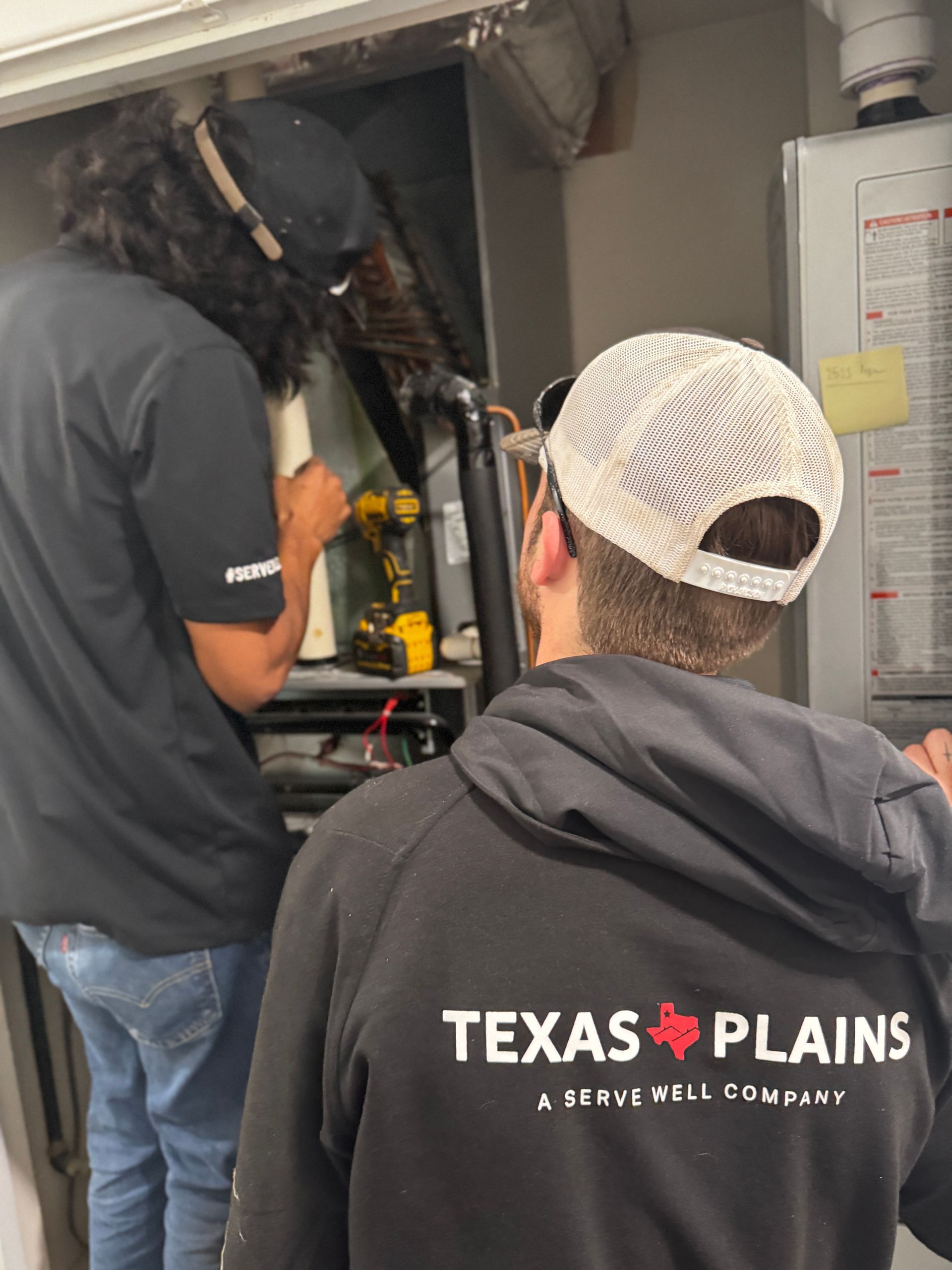 Two technicians in branded apparel work on a furnace inside a utility closet.