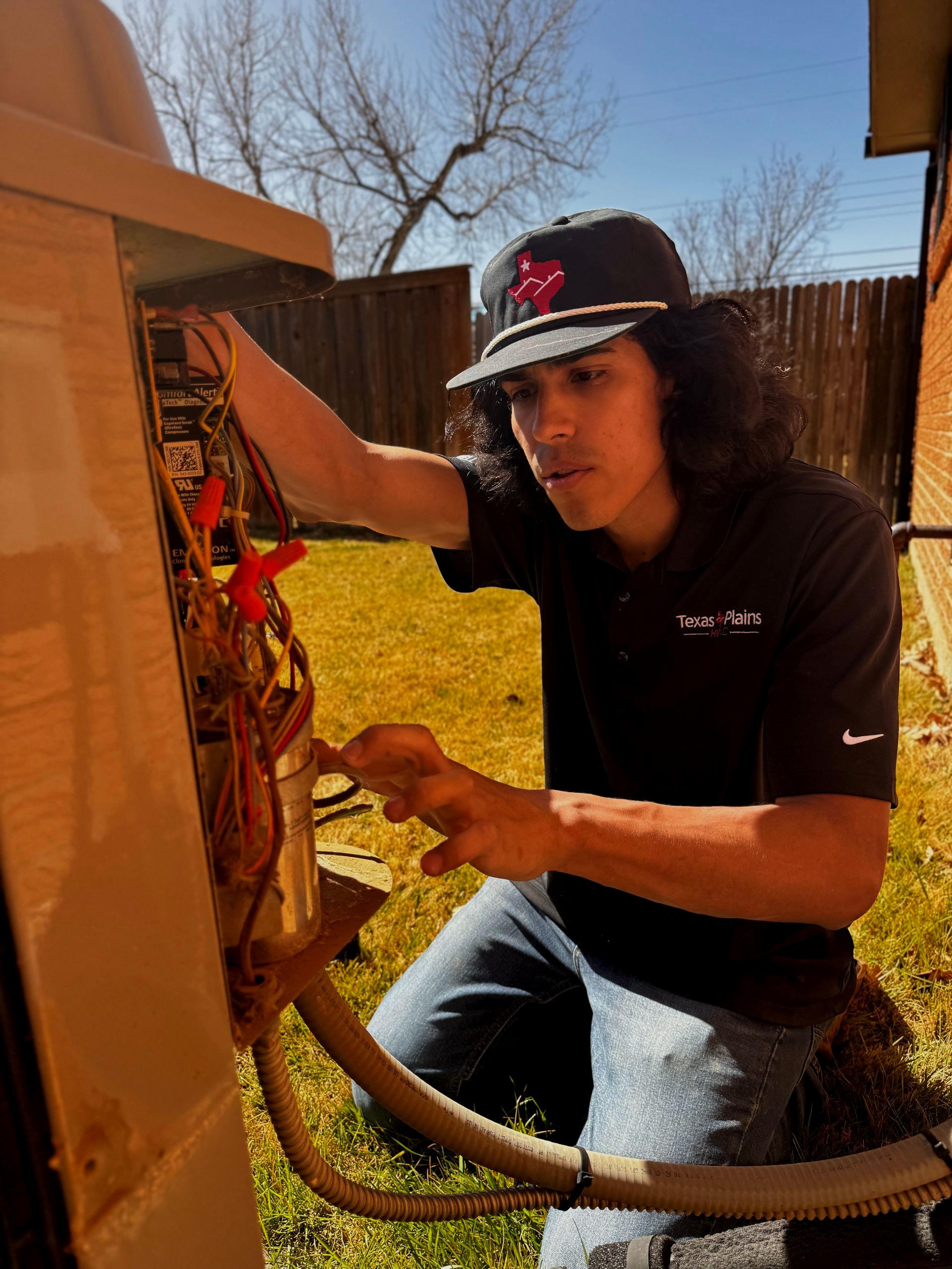 A technician kneels outdoors, repairing the wiring inside the open panel of an air conditioning unit.