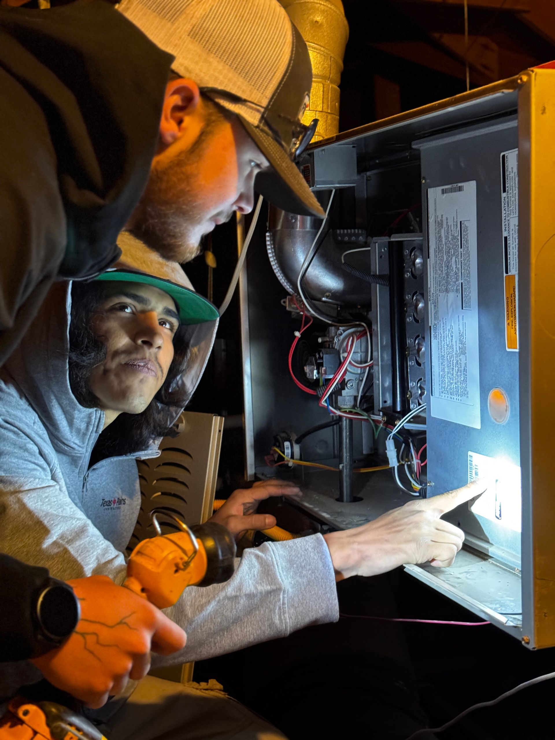 Two people examine the open electrical panel of a furnace in a dark space, one pointing to a wiring diagram.