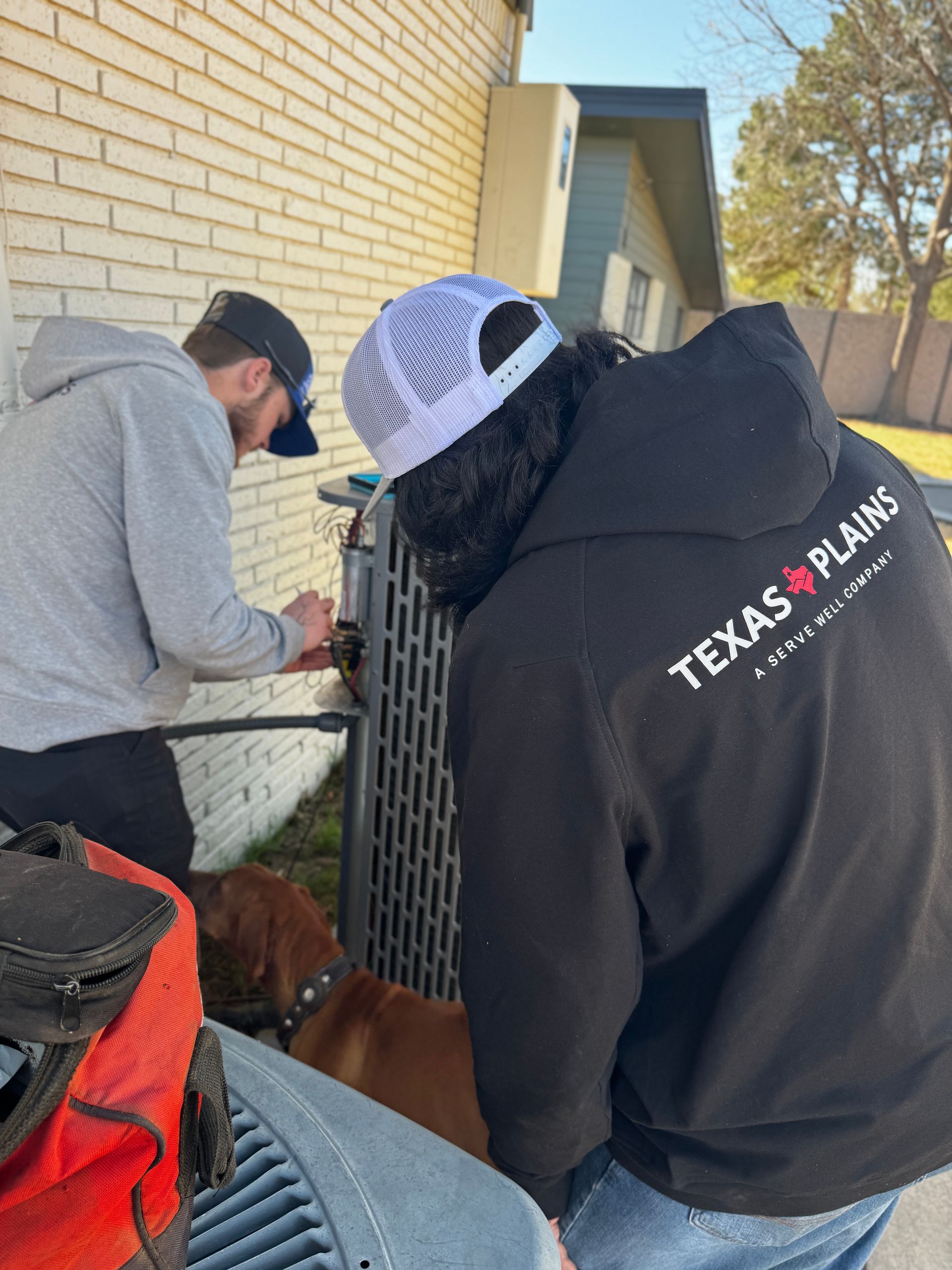 Two people work on an HVAC unit against a brick house while a brown dog stands nearby.