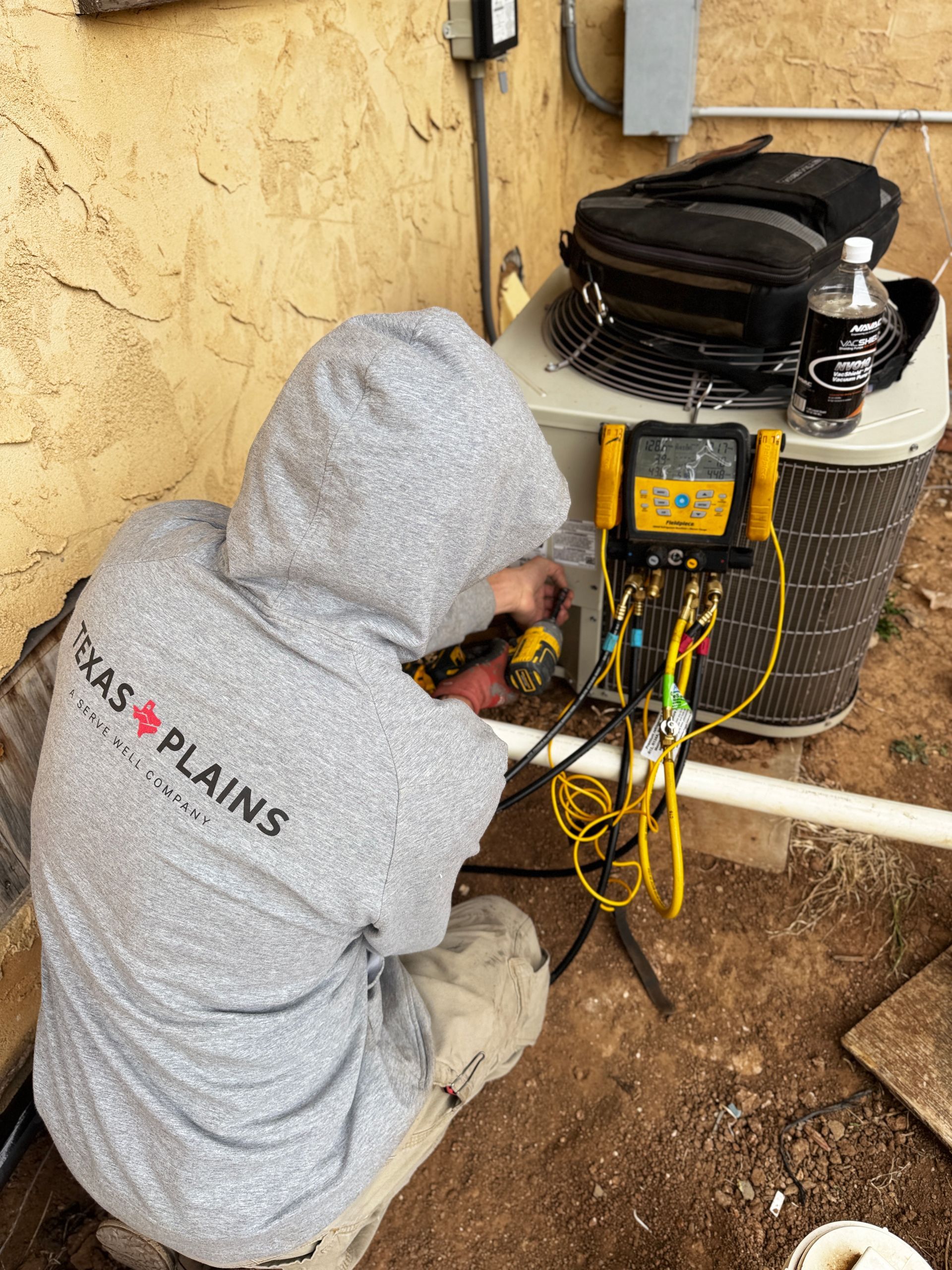 A technician in a gray hoodie works on an outdoor HVAC unit using a digital manifold gauge set and a power drill.