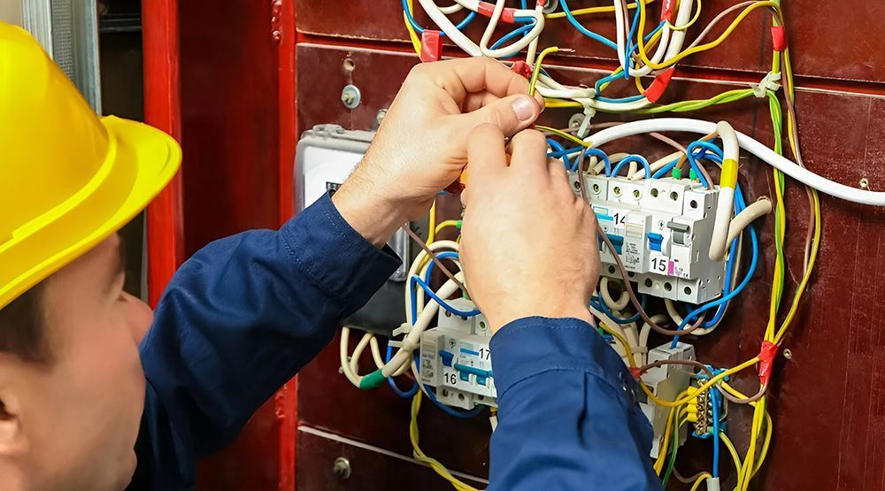 An Electrician Wearing a Yellow Hard Hat Connects Wires in a Breaker Box — Rob Ward Electrical Services in West Ballina, NSW