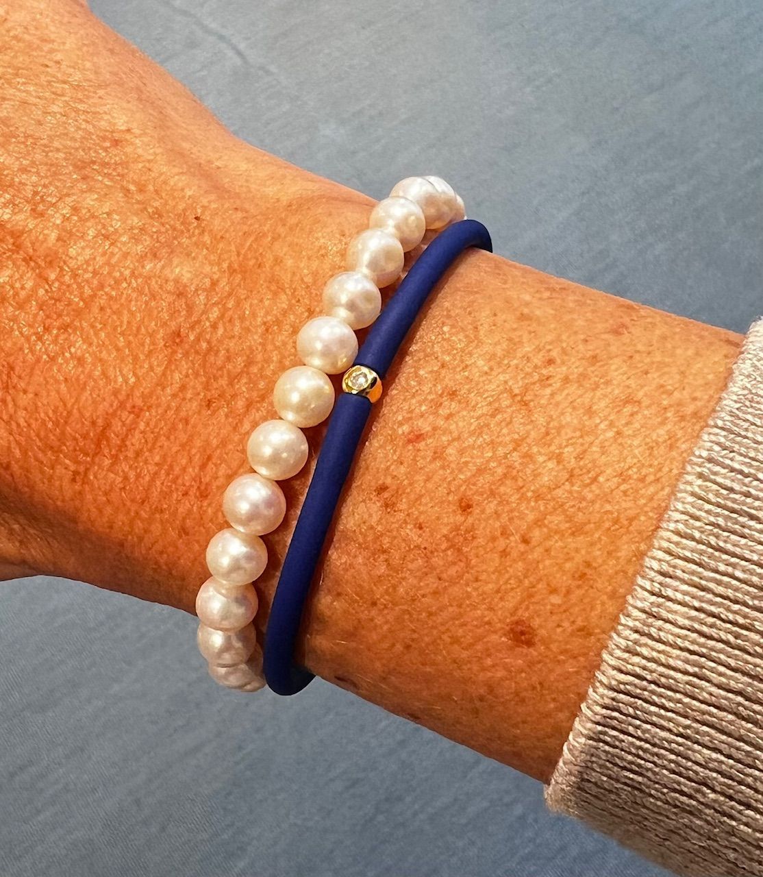 Zwei Armbänder an einem Handgelenk: eine Perlenkette und ein blaues Gummiarmband mit goldenem Akzent.