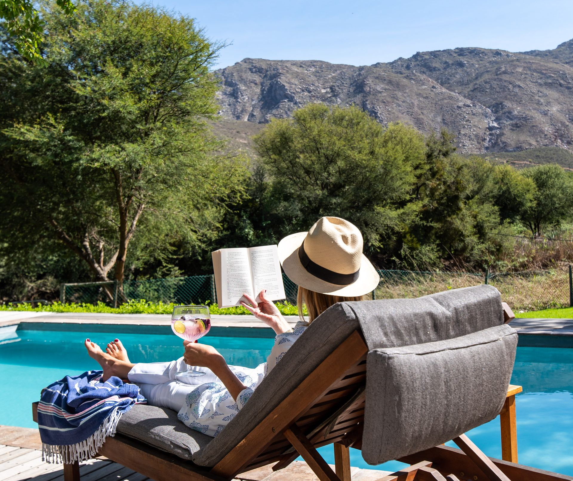 Person in straw hat relaxing by a pool, reading a book and holding a drink, with mountain backdrop.