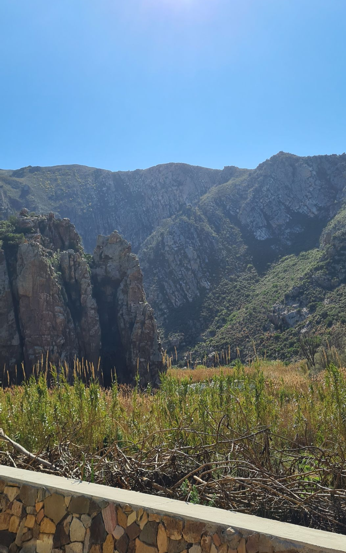 Mountains with greenery, against a blue sky, viewed from behind a low stone wall and vegetation.