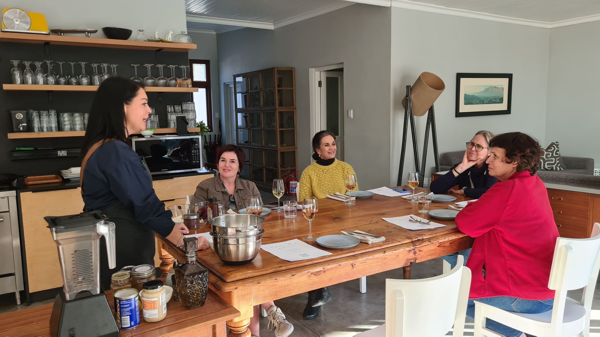 Woman presenting a wine tasting to a group seated at a wooden table in a home.